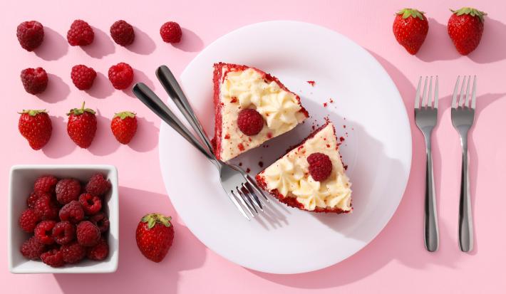 Fotografia vista de cima de um prato raso grande branco de vidro com duas fatias de torta red velvet, com dois garfos juntos ao lado. Ao redor do prato tem mais dois garfos e muitas frutas vermelhas.