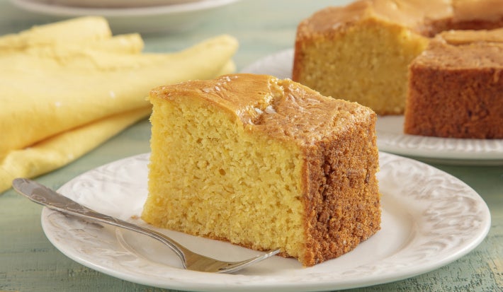 Fatia de bolo de milho macio e dourado servida em prato branco, com garfo ao lado e fundo aconchegante.