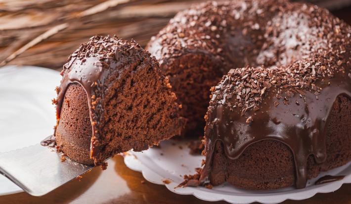 Imagem de um bolo de chocolate com cobertura de brigadeiro escorrendo e uma fatia do bolo sendo retirada com uma espátula.