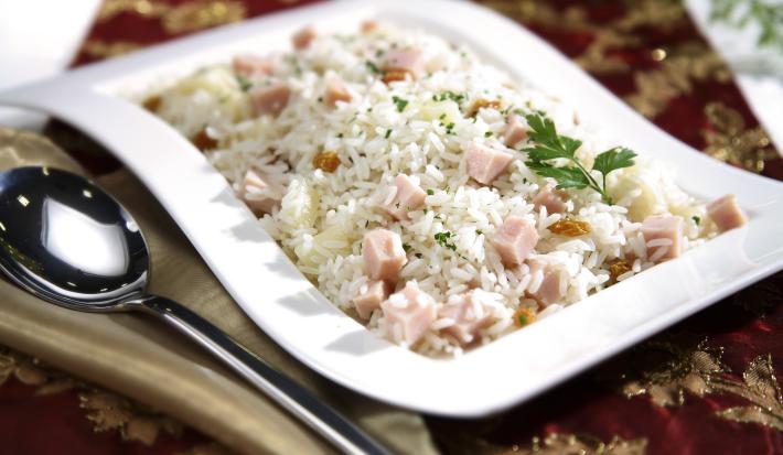 Fotografia em tons de vinho em uma mesa com uma toalha vinho e dourada, um recipiente ao centro branco com o arroz tropical com presunto e salsinha dentro dele. Ao lado, um guardanapo de pano bege com uma colher apoiada nele.