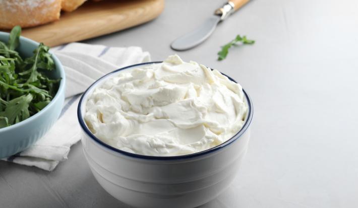 Fotografia em tons de branco com um pote branco ao centro. Dentro do pote existe uma receita de cream cheese caseiro, um creme branco feito com vinagre, leite e creme de leite.