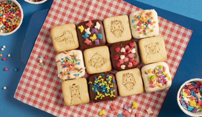 Fotografia em tons de azul e vermelho de uma bancada azul com paninho xadrez vermelho e branco sobre ele biscoitos Passatempo cobertos com chocolate e confeitos coloridos. Ao lado três recipientes brancos redondos com confeitos coloridos.