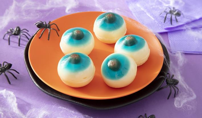 Foto em tons de laranja da receita de olhos de gelatina com Leite moça para o dia das Bruxas servida em uma tábua circular laranja sobre uma mesa roxa com aranhas de decoração