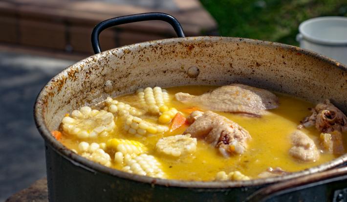 Fotografia de uma panela funda e grande com um ensopado de frango com espigas de milho e cenoura. A panela está apoiada sob alguma mesa ao ar livre.