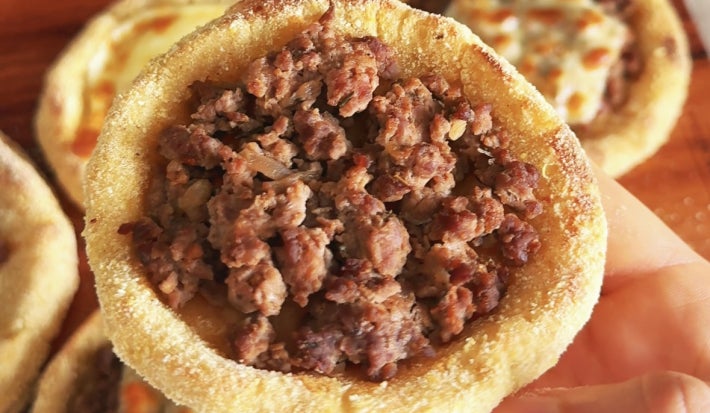 Fotografia mostra uma mão segurando uma esfiha aberta, dourada, com massa macia e levemente crocante nas bordas, recheadas com carne.