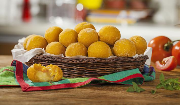 Fotografia em tons de marrom e vermelho em uma bancada de madeira com uma cesta oval de vime e vários bolinhos de abóbora assados com queijo. Ao lado, tomates inteiros e um pedaço de tomate.