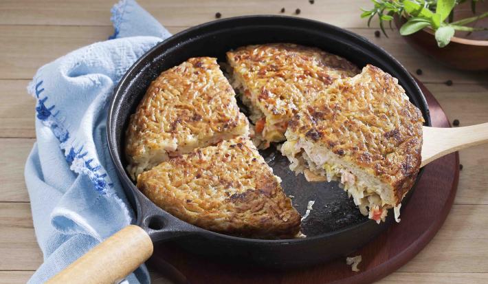 Fotografia em tons de azul em uma bancada de madeira clara com um pano azul, uma tábua de madeira escura, uma frigideira grande preta com a batata rosti recheada com legumes.