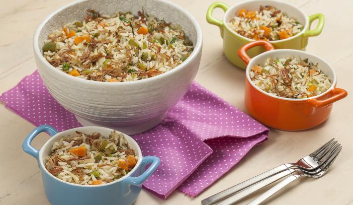 fotografia em tons de azul, laranja, branco e verde de uma bancada bege vista de cima. Contém 3 potinhos com alças nas cores azul, laranja e verde com o arroz com pedaços de carne, ao lado um recipiente redondo branco com o arroz e pedaços de carne