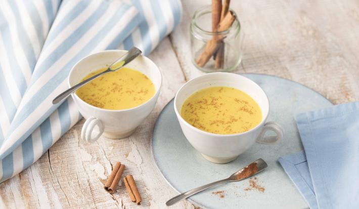 Fotografia em tons de azul em uma bancada de madeira com uma toalha branca com listras azuis, um prato raso azul com uma xícara branca em cima e a bebida de Molico Dourado dentro. Ao lado, outra xícara com a bebida e paus de canela espalhados pela mesa.