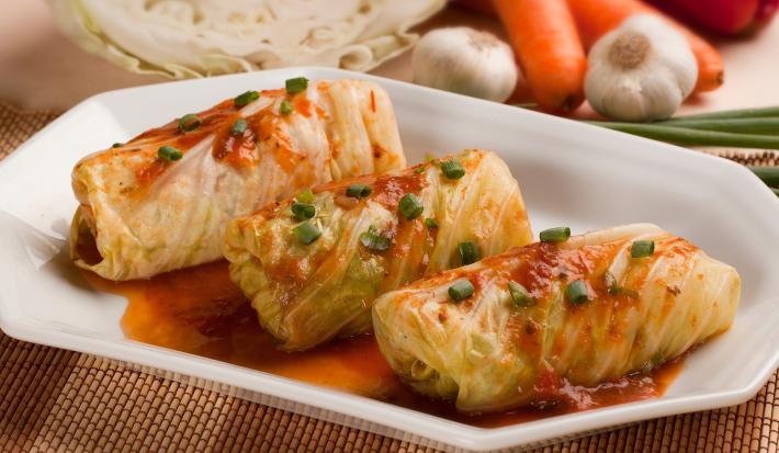 Imagem de uma travessa branca com três charutos de repolho com molho de tomate e cebolinha verde.