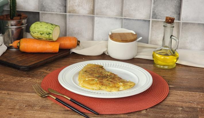 Fotografia em tons de verde e laranja em uma bancada de madeira escura com um apoiador de prato vermelho e um prato branco em cima com a crepioca de legumes. Ao fundo, abobrinha e cenoura cortadas ao meio e vidro de azeite e sal.