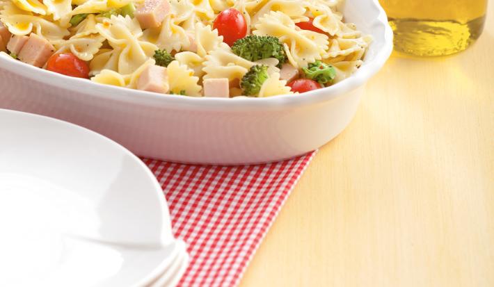 Fotografia em tons de amarelo em uma mesa de madeira com um pano quadriculado em vermelho, um recipiente oval branco com a salada de macarrão. Ao lado, um potinho com azeite.