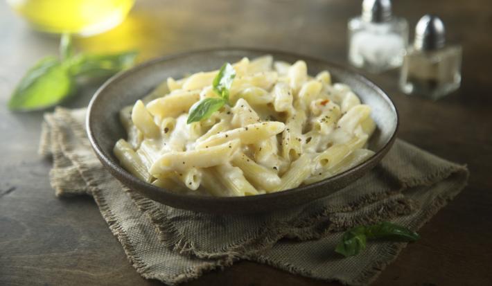 Fotografia em tons de cinza de uma bancada de madeira, ao centro um paninho cinza amarronzado com um prato redondo sobre ele com o macarrão pene com molho branco. Fotografia em tons de cinza de uma bancada de madeira, ao centro um paninho cinza amarronzado com um prato redondo sobre ele com o macarrão pene com molho branco.