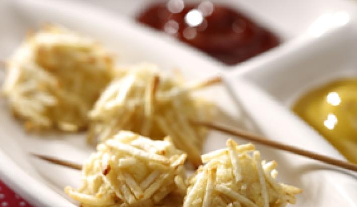 Fotografia em tons de vermelho em uma mesa com uma toalha vermelha com bolinhas brancas, um recipiente branco com o espetinho de cachorro quente empanado com batata palha, e ketchup e mostarda ao lado.