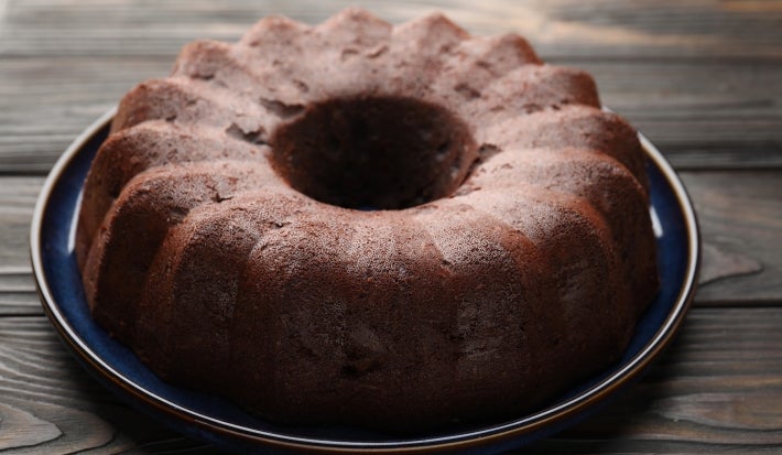 Bolo de chocolate em forma de bundt, com cor marrom escuro, está sobre um prato cerâmico azul. A superfície texturizada sugere umidade, e a apresentação rústica é complementada pela madeira do fundo.