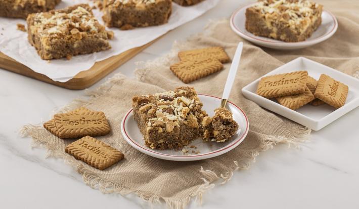 Foto da receita de Blondie de Tostines. Observa-se um prato de sobremesa com uma fatia de blondie com uma colher retirando uma fatia. Biscoitos TOSTINES decoram a foto.