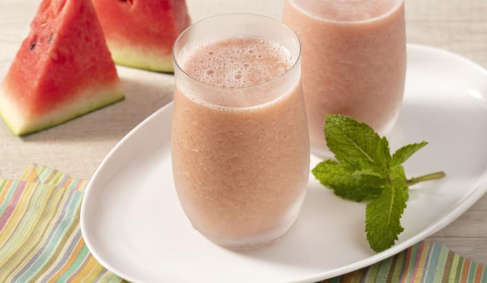Fotografia em tons avermelhados, com pedaços de melancia cortados de forma triangular, prato oval branco com folhas de hortelã e dois copos de vidro com a bebida de melancia..