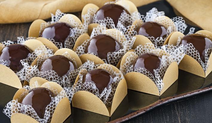 Fotografia em tons de marrom em uma bancada de madeira escura com brigadeiros caramelizados colocados em forminhas beges e ao lado um pano bege.
