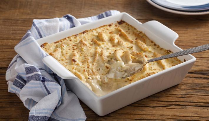 Fotografia em tons de branco e dourado, no centro um refratário com peito de frango gratinado com molho branco e uma colher de servir, ao lado, um guardanapo branco com listras em tons de azul, a borda de um prato, tudo sobre bancada de madeira rústica.