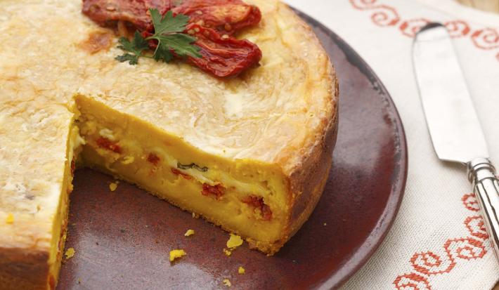 fotografia em tons de marrom e branco de uma bancada marrom vista de cima, contém um pano branco com um prato redondo marrom  com a torta faltando uma fatia e por cima tomate seco para decorar.