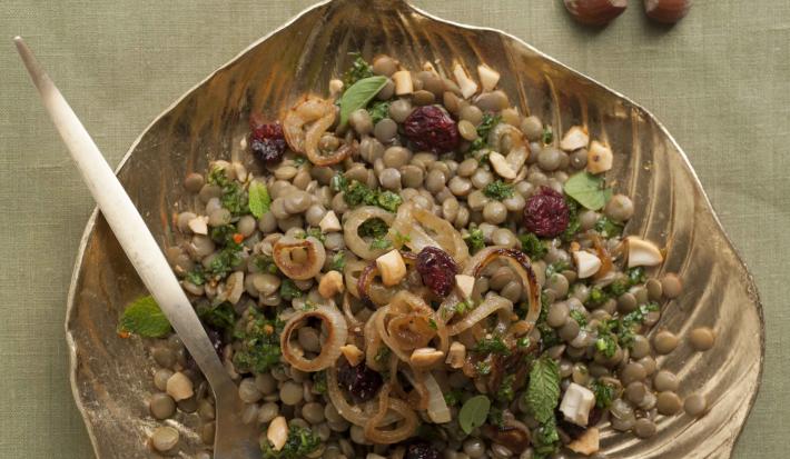 Fotografia em tons de verde em uma bancada de madeira com uma toalha verde, um prato de prata em formato de folha com a salada de lentilha, cebolas, salsa e ervas.