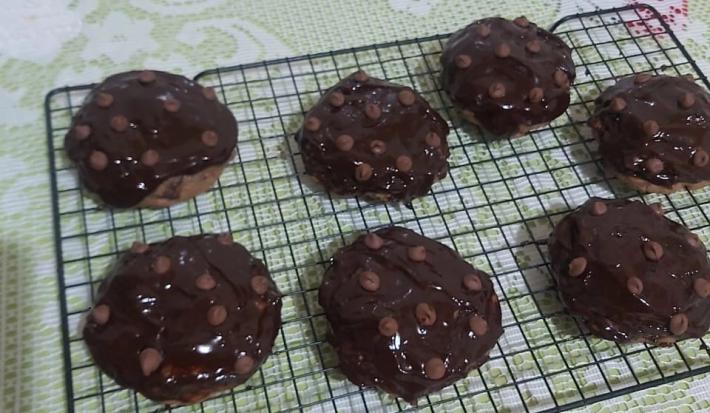 Foto da receita de Cookies low carb de chocolate. Observa-se uma grade preta com 7 cookies dispostos em cima, com calda de chocolate e chocolate em gotas na cobertura.