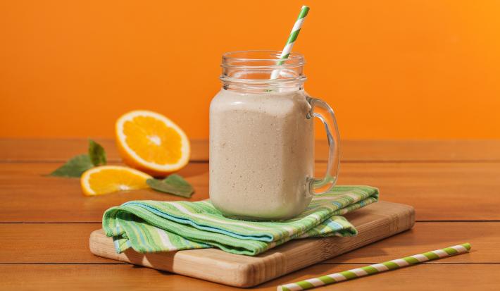 Fotografia em tons de laranja em uma bancada de madeira, um fundo laranja. Ao centro, uma tábua de madeira, um paninho verde, um canudo de papel verde e em cima um copo grande de vidro com o shake de banana dentro. Ao fundo, uma laranja aberta e folhas.