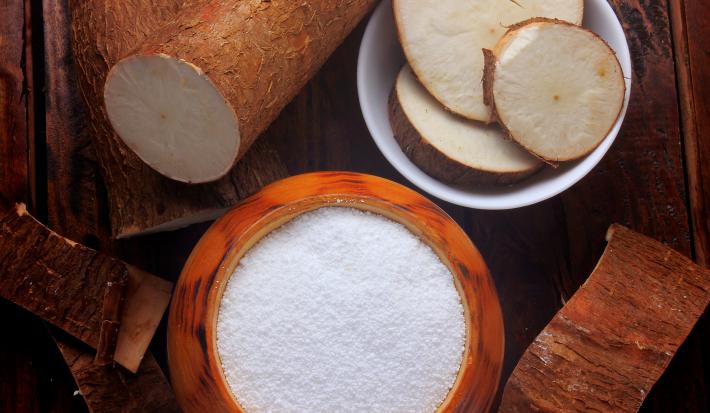 fotografia em tons de marrom e branco de uma bancada marrom vista de cima, contém um recipiente redondo marrom com a farinha dentro e ao lado um recipiente redondo branco com pedaços de mandioca. Em volta contém mandiocas.