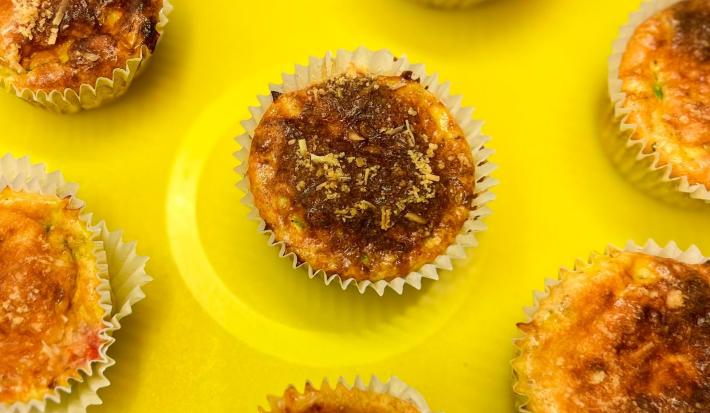 Fotografia em tons de amarelo com vários muffins de omelete ao centro. Em cima de cada muffin existe uma camada de queijo parmesão