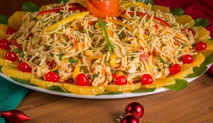 Em cima de uma mesa, prato de salpição colorido com frango desfiado, pimentões em tiras, milho-verde e batata-palha, decorado com laranja em rodelas, tomates e no topo do prato fio de cenoura em forma de flor.