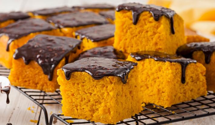 Fotografia em tons de amarelo e preto de uma bancada de madeira com um potinho branco com calda de chocolate, ao lado uma grade preta com fatias quadradas de bolo de cenoura com calda de chocolate em cima, ao fundo, um pano amarelo.