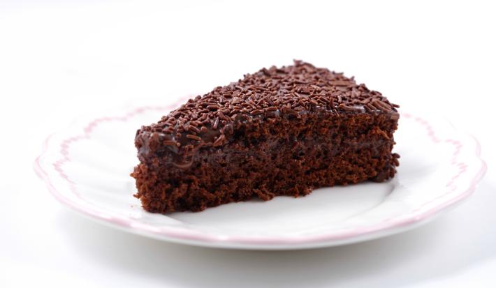 Imagem de uma fatia de bolo de chocolate com recheio e cobertura de brigadeiro e chocolate granulado sobre um prato branco.