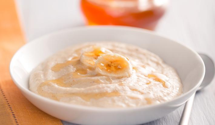 Fotografia em tons de branco, cinza e laranja, com prato branco ao centro contendo um creme decorado com fatias de banana e mel, no entorno guardanapo laranja, potinho com mel e uma colher, tudo sobre bancada em tons de cinza.