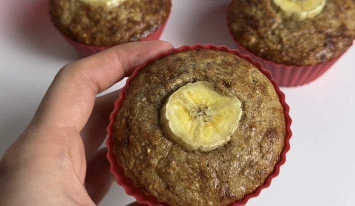 Fotografia de uma mão segurando um muffin de banana com aviea em uma forminha de silicone. No fundo tem mais muffins sobre uma mesa branca.