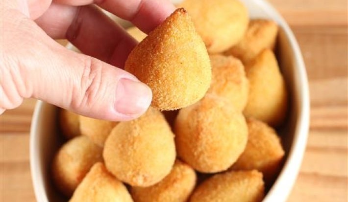 Fotografia de uma mão segurando uma coxinha pequena. Ao fundo tem recipiente branco de vidro oval sobre uma mesa de madeira. Dentro do recipiente tem várias mini coxinhas.