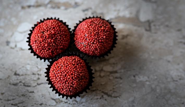 Fotografia em tons de vermelho, em uma bancada de madeira de cor cinza. Ao centro, 3 formilhas de brigadeiro contendo os brigadeiros.