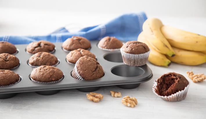 Fotografia em tons de cinza, marrom e amarelo, forma com cupcakes de chocolate em bancada cinza, nozes espalhadas ao redor, ao lado um cacho de banana e um cupcake aberto e ao fundo um guardanapo azul com listras brancas.