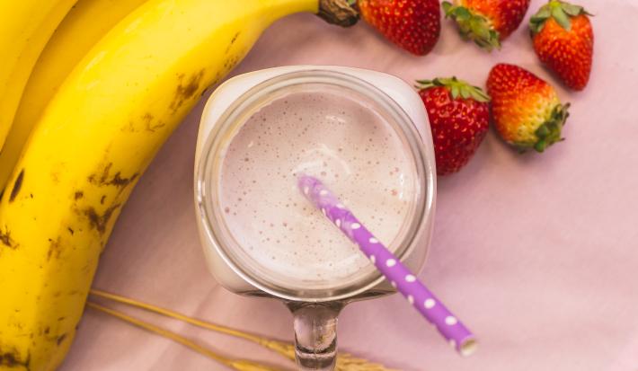 Fotografia em tons de rosa, de uma bancada rosa vista de cima, ao centro um copo com uma bebida e um canudo, em volta bananas e morangos.