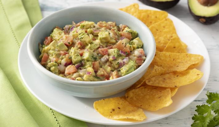 Fotografia em tons de branco e verde de uma bancada de madeira branca com um paninho verde e um prato redondo branco, sobre ele um recipiente branco redondo com a guacamole e nachos. Ao fundo um avocado e folhas de salsinha.