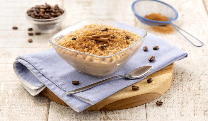 Fotografia em tons de azul em uma bancada de madeira clara, uma tábua de madeira redonda ao centro com um paninho azul. Em cima, um pote de vidro com o arroz doce dentro polvilhado com canela em pó e decoração com canela em pau.