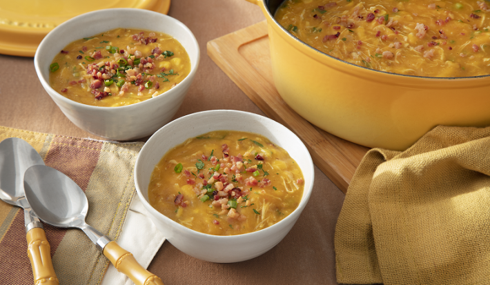 Foto em tons de amarelo da receita de caldo de kenga servida em dois bowls brancos de porcelana com duas colheres prateadas ao lado. Ao lado uma panela com o caldo e um paninho, ambos amarelos, sobre uma tábua de madeira. Ao fundo, a tampa da panela