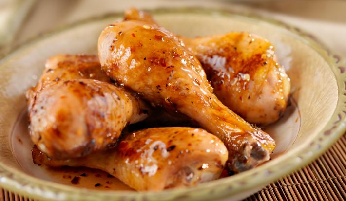 Fotografia de cinco coxas de frango refogadas com um brilho do caldo dentro de um recipiente fundo de vidro de cor clara, que está sobre um apoio de mesa com decoração de madeira.