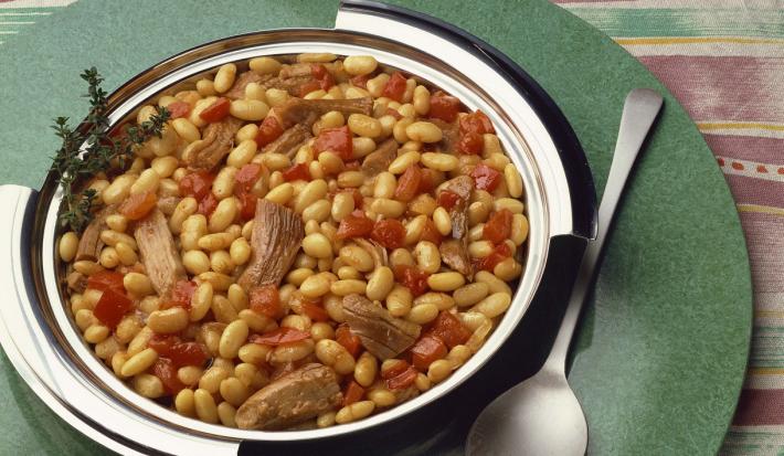 Fotografia em tons de verde em uma bancada de madeira listrada. Ao centro, uma tábua redonda e verde contendo um prato de inox com o cassoulet. Ao lado, há uma colher.