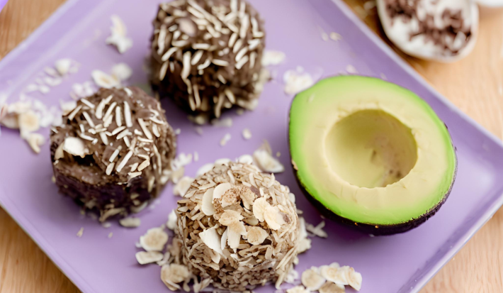 Fotografia em tons de roxo com um prato retangular roxo ao centro. Em cima do prato existe 3 brigadeiros de cor marrom e ao lado existe metade de um abacate. Todos os brigadeiros estão polvilhados com coco ralado