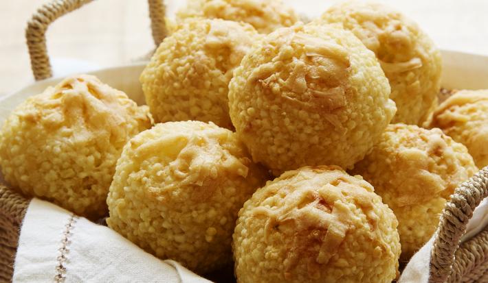 Fotografia em tons de marrom em uma bancada de madeira com uma cestinha de vime com vários pãezinhos de tapioca redondos dentro.