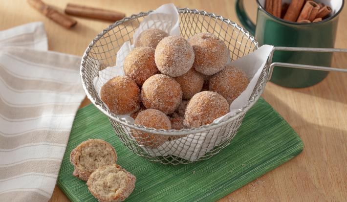 Foto da receita de bolinho de chuva sem glúten. Observa-se uma peneira com os bolinhos dentro, polvilhados de açúcar e canela, sobre um guardanapo de pano verde