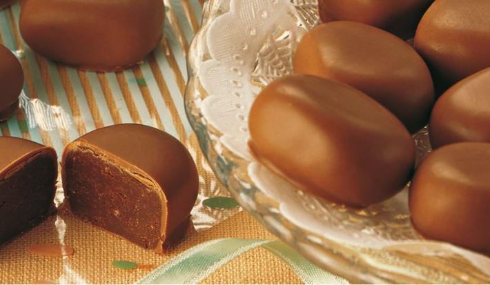 Fotografia em tons de marrom, em uma bancada de madeira com um suporte para bolo de vidro e vários bombons de chocolate em cima dele.