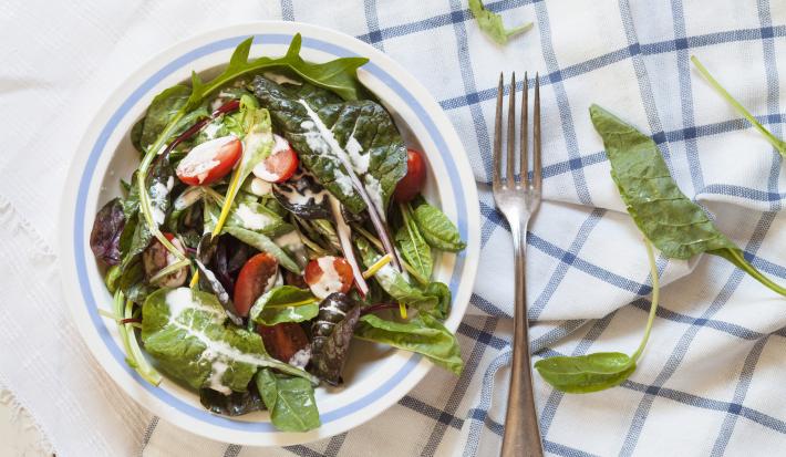 Fotografia vista de cima de um recipiente fundo branco com uma listra azul claro na borda, com uma salada de folhas, com molho de creme de leite e tomates cereja. O recipiente está sobre duas toalhas de mesa, nas cores branco e azul, e ao lado, um garfo.