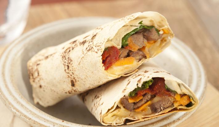 Fotografia em tons de marrom em uma bancada de madeira com uma tábua de madeira clara, um prato redondo fundo com dois wraps de carne com legumes.