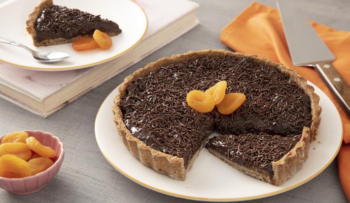 Foto da receita de Torta de chocolate com damasco. Observa-se a torta em um prato grande branco, com granulados em cima e três damascos decorando. Para trás, ao lado esquerdo, tem um prato de sobremesa branco com uma faia da torta, sobre um livro.
